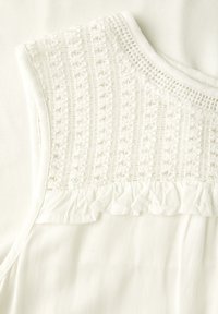 Weiße Bluse mit strukturierten, gestickten Details am Ausschnitt, weichem Stoff und einem rüschend verzierenden Akzent am unteren Rand.
