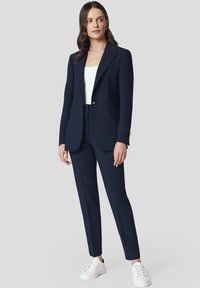 Granatowa marynarka o dopasowanym kroju z pojedynczym złotym guzikiem, zestawiona z dopasowanymi spodniami i białymi trampkami; gładki materiał o prostym wzornictwie.