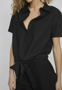 Čierna košeľa s krátkymi rukávmi a golierom, má zapínanie na gombíky vpredu a uviazaný pás. Vyrobená z mäkkej, štruktúrovanej látky.