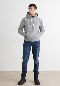 Felpa grigia con cappuccio e coulisse, indossata sopra una maglietta nera, abbinata a jeans blu e stivaletti neri. Design semplice con un piccolo logo sul petto.