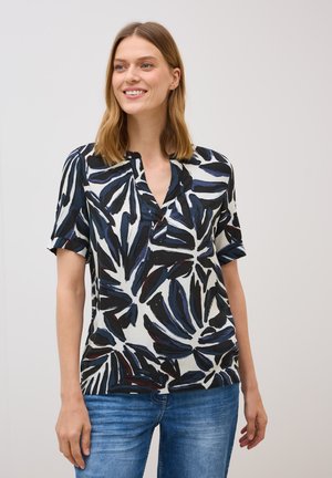 Femme souriante portant une chemise à motifs avec des dessins de feuilles noirs et bleus, vêtue de jeans, sur fond blanc uni.