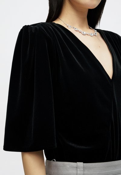 Μαύρη βελούδινη μπλούζα με κοντά φουσκωτά μανίκια και βαθύ V-ντεκολτέ. Διακοσμημένη με ασημένια κολιέ που διαθέτει πολλαπλούς λαμπερούς λίθους.