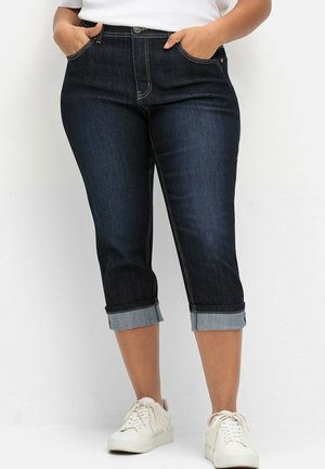Mørk denim capri bukser med opfoldede manchetter, der har forlommer og en klassisk pasform. Båret med hvide sneakers på en neutral baggrund.