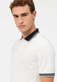 Weißes Poloshirt mit kurzen Ärmeln, zwei Knopfverschluss, marineblauem Kragen mit mehrfarbigen Streifen und einem dezenten Logo auf der Brust.