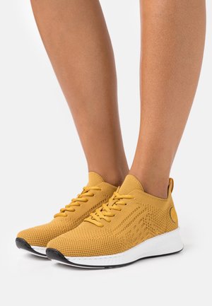 Żółte musztardowe dzianinowe trampki z białymi podeszwami na bosych stopach, pokazane z boku i z przodu na jednolitym białym tle.