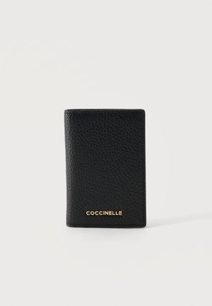 Sort tekstureret læderpung stående oprejst med guld "COCCINELLE" logo centreret nær bunden på en ensfarvet hvid baggrund.