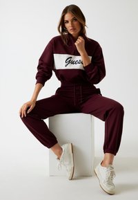 Felpa bordeaux con pannello bianco che presenta il logo "Guess", abbinata a pantaloni joggers coordinati e sneakers bianche. Tessuto morbido, vestibilità rilassata.