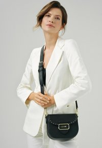 Czarna skórzana torba na ramię o zaokrąglonym kształcie, z ozdobami w formie ćwieków, złotymi elementami i regulowanym paskiem. Na neutralnym tle.