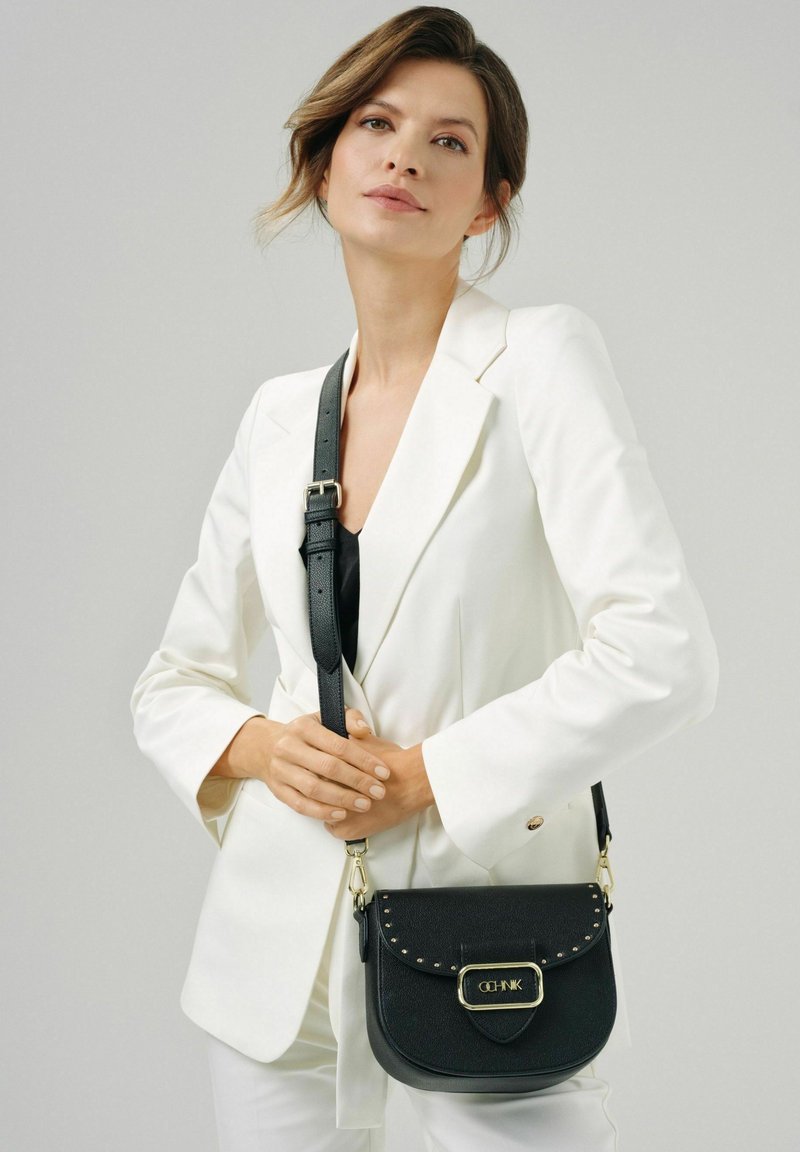 Czarna skórzana torba na ramię o zaokrąglonym kształcie, z ozdobami w formie ćwieków, złotymi elementami i regulowanym paskiem. Na neutralnym tle.