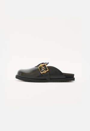 Juodos odinės slip-on tipo bateliai su auksine sagtimi, apvaliu nosiu ir tekstūrine guma padulkiu. Paprastas dizainas su atvira kulna.