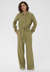 Femme avec les cheveux courts bruns portant une combinaison olive à boutons avec ceinture et pantalons larges, debout les mains dans les poches.
