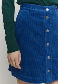 Blaue Cord-Rock mit vertikalen Rippungen, Knopfverschluss vorne, zwei Seitentaschen und einem leicht ausgestellten Saum. Texturierte Stoffdetails sichtbar.