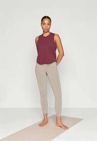 Maroon sleeveless top paired with beige legging pants. Thin, lightweight fabric. Model stands on a beige yoga mat. Simple design.