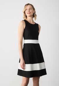 Svart A-linje klänning med en figurnära bodice och breda vita ränder vid midjan och nederkanten. Tillverkad av ett slätt tyg med en klassisk, ärmlös design.