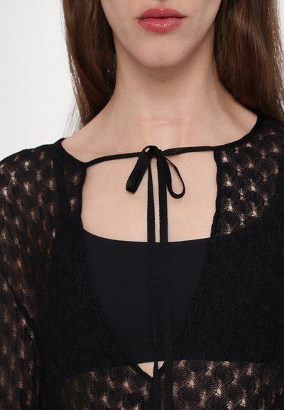 Top in pizzo nero con un design a rete decorato e chiusura a cravatta sul collo, sovrapposto a un tessuto nero sottostante.