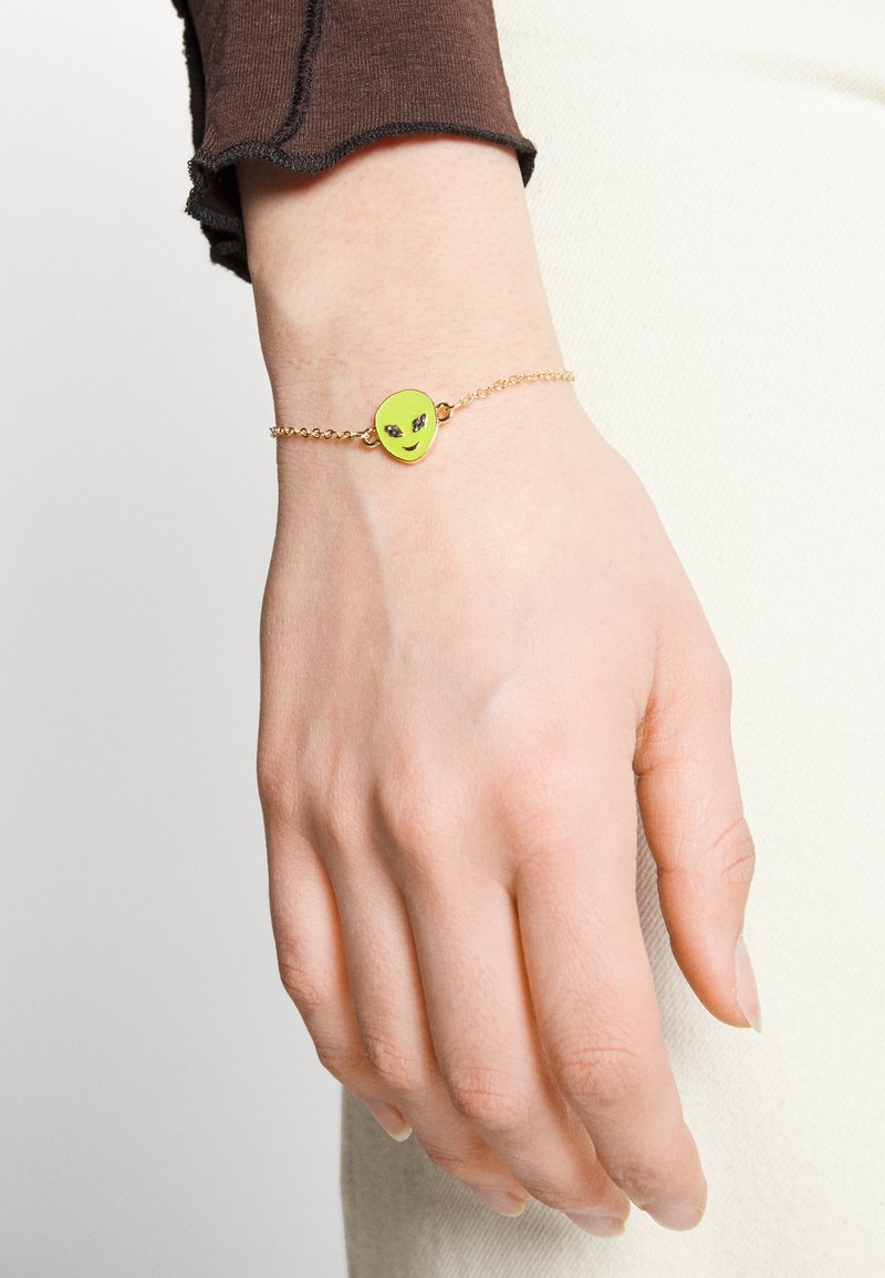 Pulseira de corrente dourada com um charme de rosto de alienígena verde com uma expressão sorridente e detalhes em prata para os olhos.