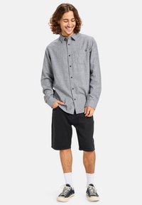 Gray flannel shirt with a button-down front and pocket, paired with black shorts. Model wears white socks and black sneakers. Casual fit.