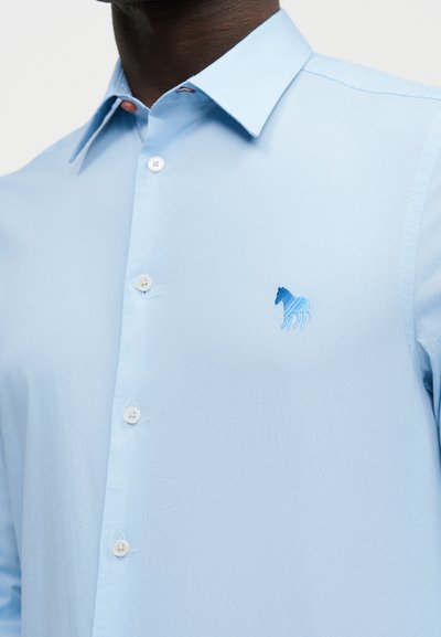 Άνδρας που φοράει ανοιχτό μπλε πουκάμισο με κουμπιά και έχει ένα μικρό μπλε λογότυπο αλόγου κεντημένο στο αριστερό μέρος του στήθους.