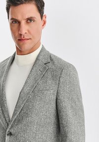 Jasnoszary marynarka w jodełkę z klapami, pojedynczym guzikiem i kieszenią na piersi. Założona na bawelniany sweter z wysokim kołnierzem w kremowym kolorze.