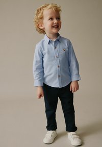 Cămașă albastru deschis cu mâneci lungi, nasturi maro și o broderie mică cu un animal, asortată cu pantaloni bleumarin și adidași albi.