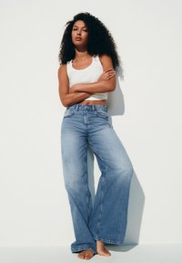 Donna con capelli ricci che indossa una canotta bianca e jeans blu a gamba larga, in piedi scalza contro un muro bianco.