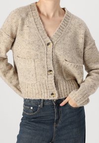 Osoba ubrana w beżowy sweter zapinany na guziki z przednimi kieszeniami oraz niebieskie spodnie z wysokim stanem, z jedną ręką w kieszeni.