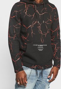 Zwarte hoodie met een rood bliksempatroon, met een tekst op de voorkant die "REBEL" en een citaat bevat, gedragen met lichtblauwe distressed jeans.
