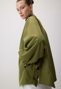 Oliwkowa, obszerna kurtka o gładkiej fakturze, z falbankowymi detalami po bokach i kieszeniami bocznymi. Model stoi na jasnym tle.