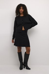 Černý pletený cardigan s knoflíky a zkráceným designem, kombinovaný s černou upnutou sukní. Modelka má na sobě černé kožené boty po kolena.
