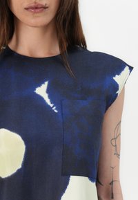 Mørkeblå topp med rund hals, kortermede ermer og en lomme på brystet. Stoffet har et strukturert, abstrakt design med lyse kremfargede detaljer.