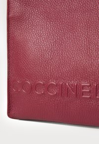 Burgundy leather wallet featuring a textured finish. Embossed "COCCINELLE" logo at the bottom, with clean edges and a compact design.