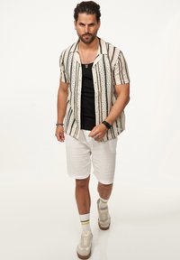 Kurzärmliges Hemd in Beige mit schwarzen vertikalen Streifen und Mustern, kombiniert mit weißen Shorts und Sneakers mit hellen Akzenten.