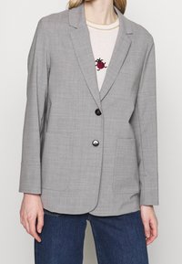 Šedý blazer s klasickým límcem, dvěma předními kapsami, zapínáním na jeden knoflík a texturovaným materiálem. Nosený přes světlý svetr.