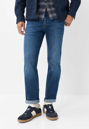 Mand iført opkrøllede blå jeans, hvide sokker og sort-hvide sneakers, med en del af en ternet skjorte og navy jakke synlig.
