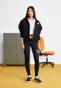 Čierna zipsová mikina, biely krátky tričko, čierne legíny s logom a čierne tenisky so žltými pruhmi. Oranžová stolička v pozadí.