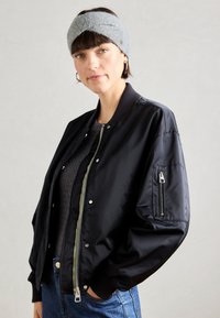 Black bomber jacket with ribbed cuffs and collar, zippered front, side pockets, and an additional zip pocket on the sleeve. Textured dark top.