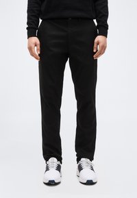 Άνδρας που φοράει μαύρο παντελόνι και λευκά αθλητικά παπούτσια, στέκεται μπροστά σε ένα απλό λευκό φόντο, τα χέρια χαλαρά στα πλευρά.