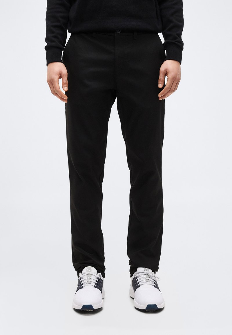 Άνδρας που φοράει μαύρο παντελόνι και λευκά αθλητικά παπούτσια, στέκεται μπροστά σε ένα απλό λευκό φόντο, τα χέρια χαλαρά στα πλευρά.