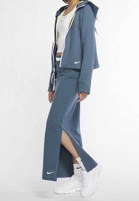 Sweat à capuche zippé bleu avec un motif texturé, superposé à un débardeur blanc, associé à une jupe longue bleue assortie avec des fentes sur les côtés et des baskets blanches.