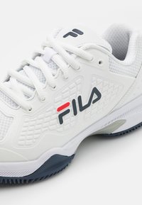 Adidași albi de tenis cu toc sintetic texturat, design cu șireturi, logo Fila în gri și roșu, talpă din cauciuc bleumarin și guler pentru gleznă căptușit.