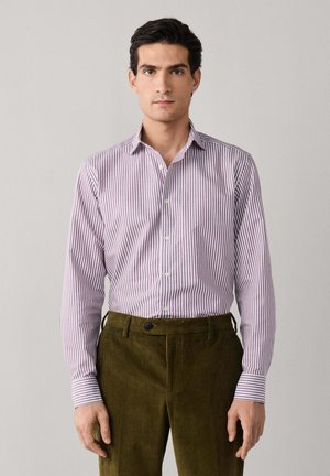 Hombre con una camisa ajustada de manga larga con rayas moradas y blancas abotonada y pantalones de pana verde oliva, de pie contra un fondo liso.