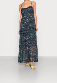 Floral maxi dress in dark navy with adjustable spaghetti straps, layered ruffles, and delicate patterns; paired with lime green block-heeled sandals.