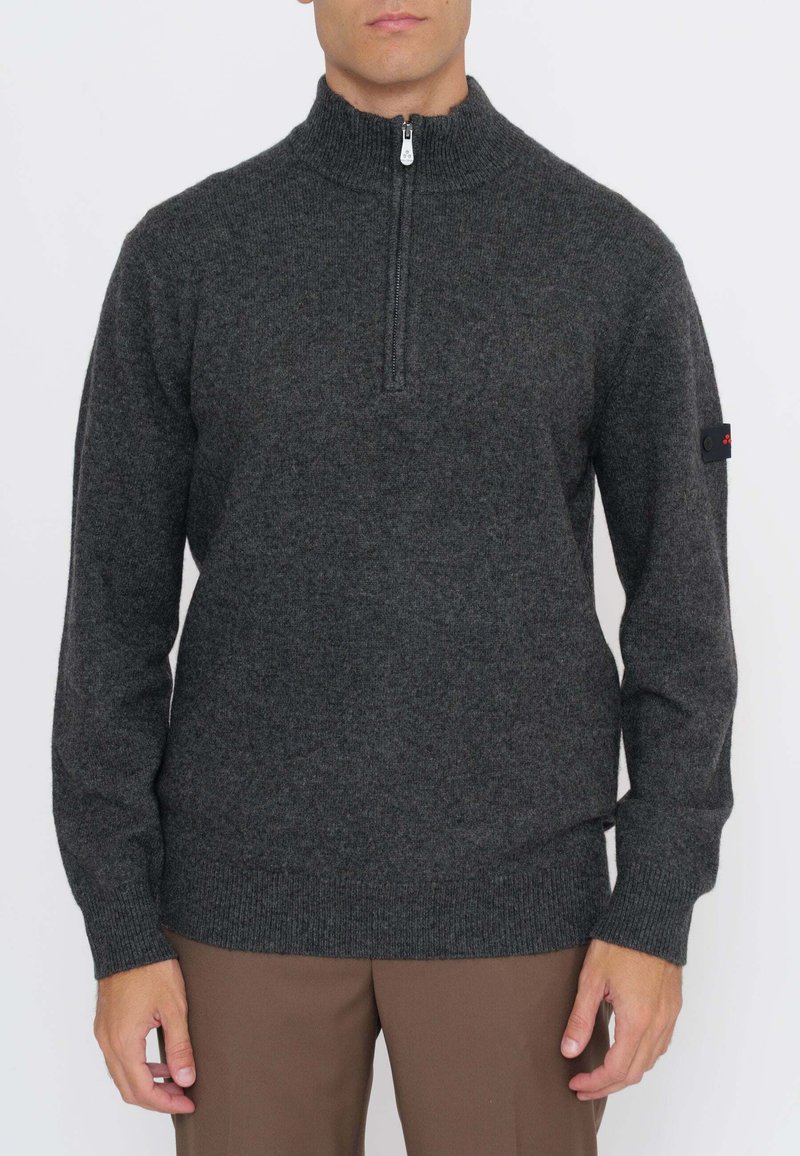 Uomo che indossa un maglione grigio scuro con mezza cerniera, polsini e collo a coste, abbinato a pantaloni marroni, in piedi su sfondo bianco.