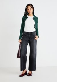 Cardigan verde con estampado sobre una blusa blanca de canalé, combinado con pantalones oscuros de pierna ancha y zapatos patentes burdeos. Sosteniendo un pequeño bolso burdeos.
