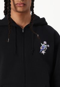 Svart glidelåshettegenser med frontlomme, prydet med et fargerikt brodert grafisk motiv av en figur og stjerner på venstre brystområde.