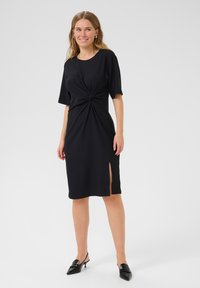 Robe noire jusqu'aux genoux avec des manches courtes, design à l'avant tordu, fente sur le côté et tissu lisse et texturé. Portez-la avec des talons slingback noirs.