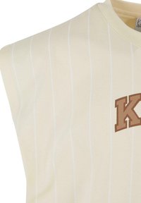 Šviesiai geltonas be rankovių marškinėlis, turintis plonas baltas vertikalias juosteles ir rudą "K" logotipą priekyje. Minkšta audinio tekstūra.
