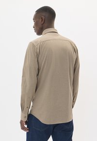 Šviesiai ruda ilgo rankove marškiniai, pagaminti iš tekstūruotos medžiagos, su standartiniu apykaklė ir mygtukais užsegamomis rankovėmis, matomi iš užpakalio.