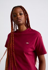 Noor must ikoon tume nahk ja pikad punased punutised, seljas marooni värvi Dickies t-särk, üksluisel heledal taustal.