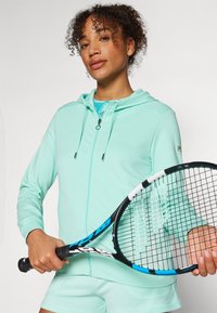 Felpa verde menta con zip, cappuccio e tasche, indossata sopra una maglietta azzurra. La persona tiene una racchetta da tennis con accenti neri e blu.
